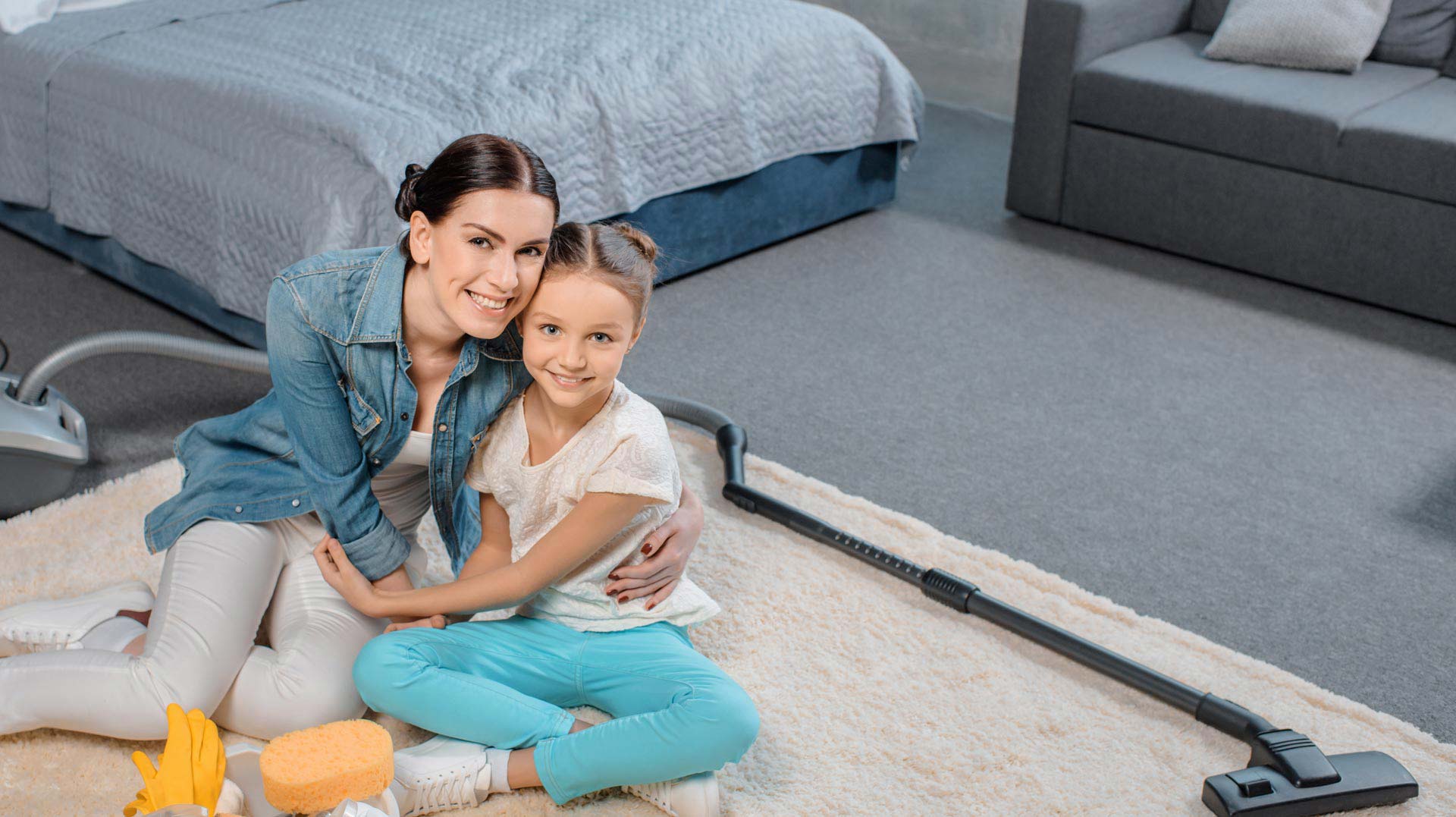 Carpet Clean Team: Mother and daughter smiling at the camera while sitting on a carpeted floor with cleaning supplies and a vacuum cleaner, in a tidy room.